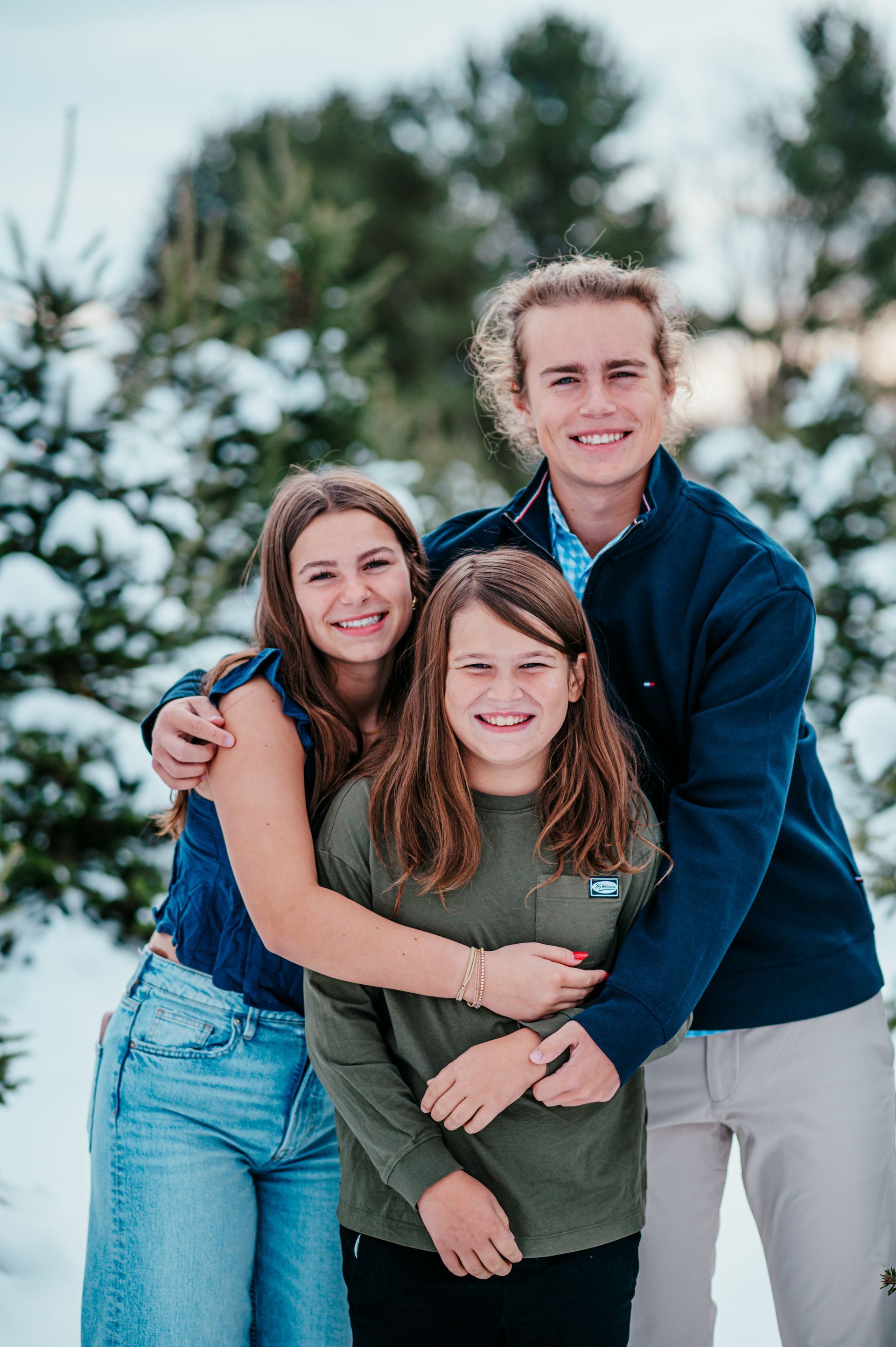 Three friends smiling and hugging outdoors in winter snow, with trees in the background