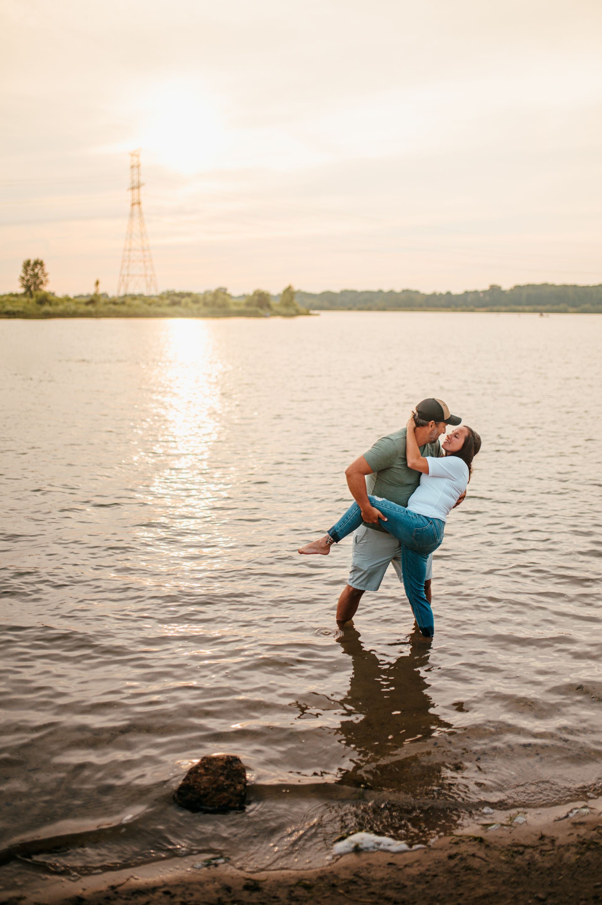 Couple embracing in a shallow lake at sunset, with calm water and a distant shoreline.