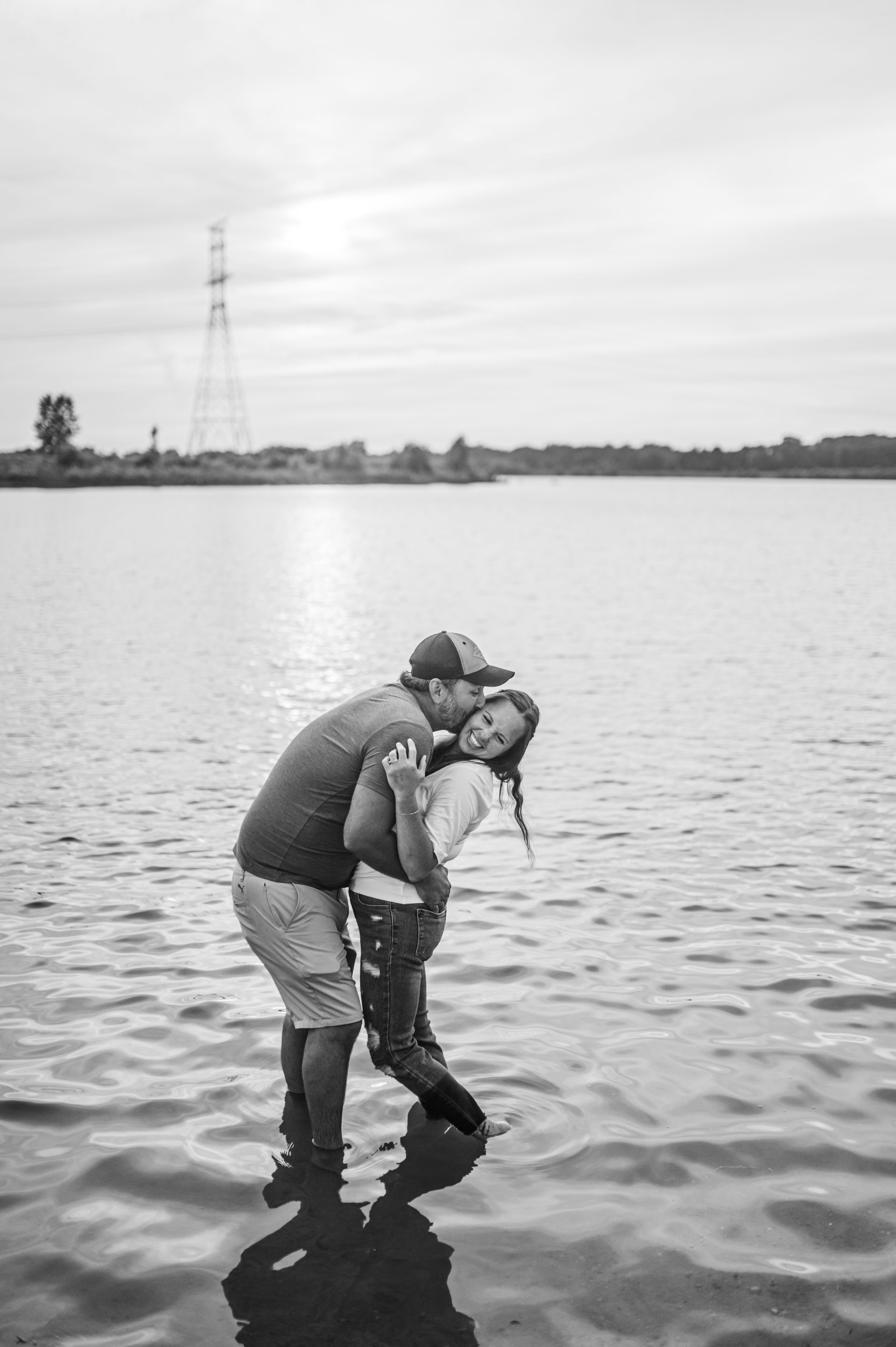 Person carrying another person through shallow water, with a calm lake and cloudy sky in the background.