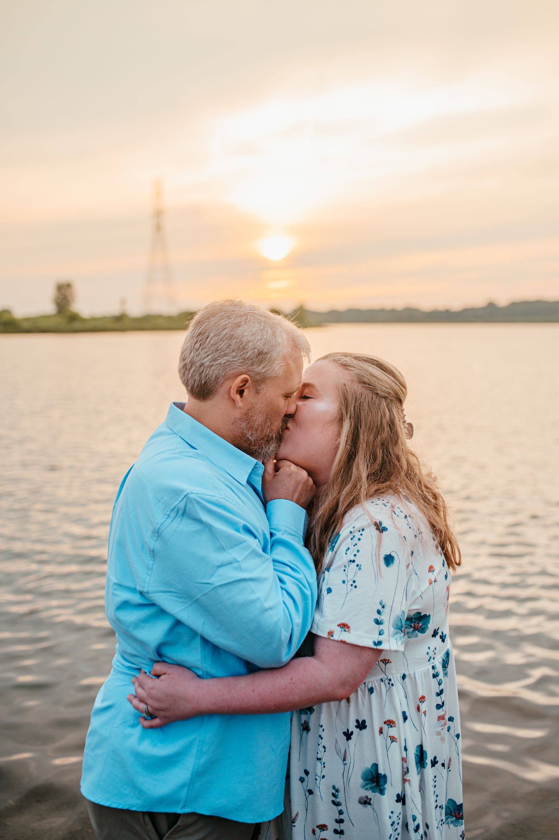 Couple embracing and kissing by a lake at sunset, with warm light and calm water.
