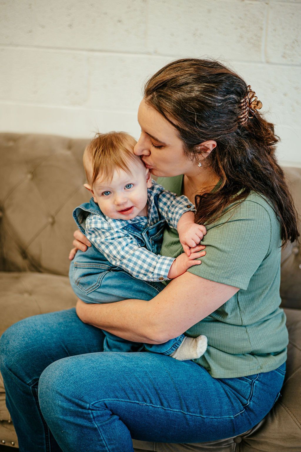Woman holding and kissing a smiling toddler on a beige couch, both wearing blue clothing.