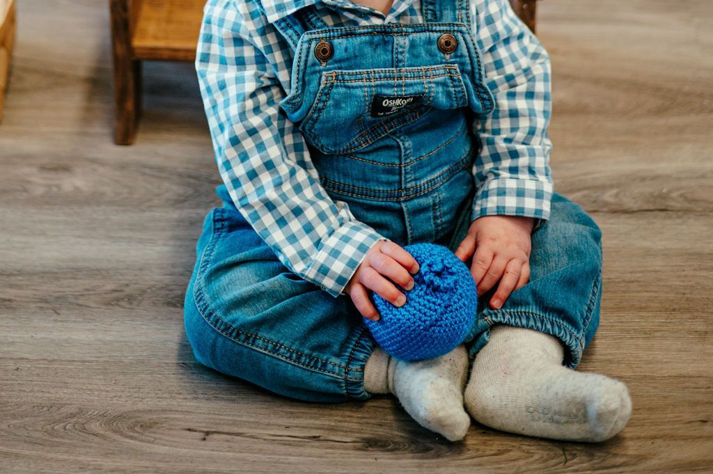 Child in denim overalls sitting on a wooden floor, holding a blue textured ball.