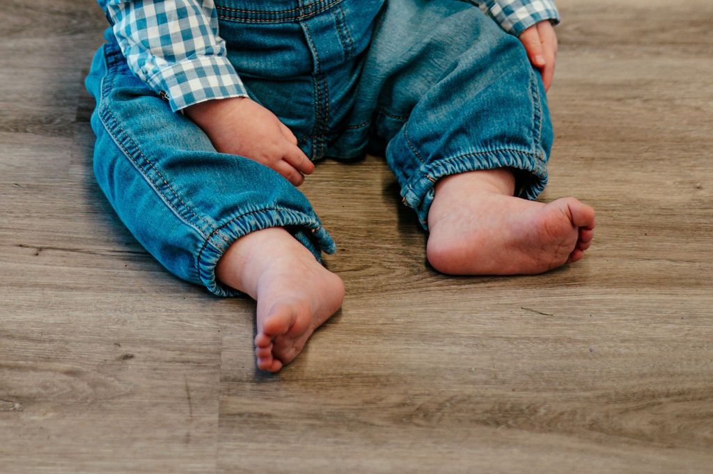 Child in blue jeans and plaid shirt sitting barefoot on a wooden floor, legs stretched out.