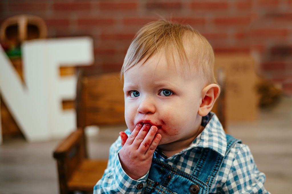 Toddler in blue plaid overalls with hand near mouth, seated indoors against a brick wall background