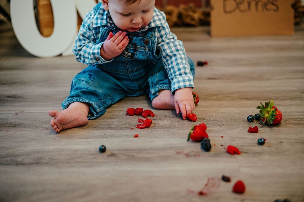Baby in blue denim overalls sitting on a wooden floor with scattered strawberries and blueberries