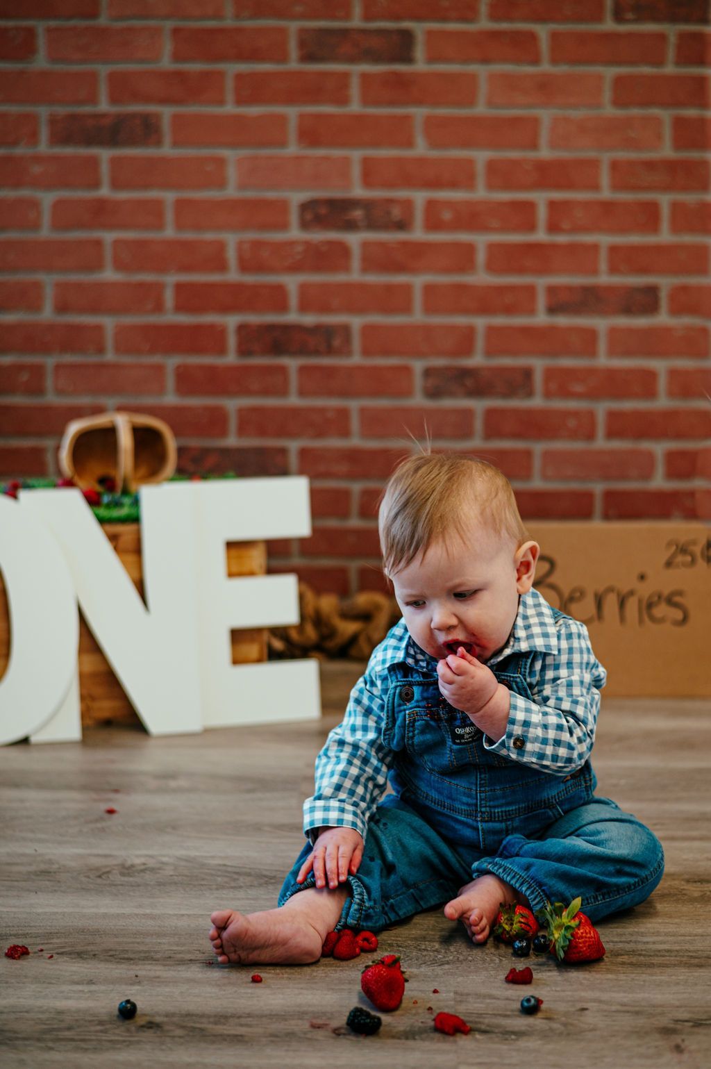 Child in blue overalls sitting on a wooden floor, examining strawberries with a brick wall background