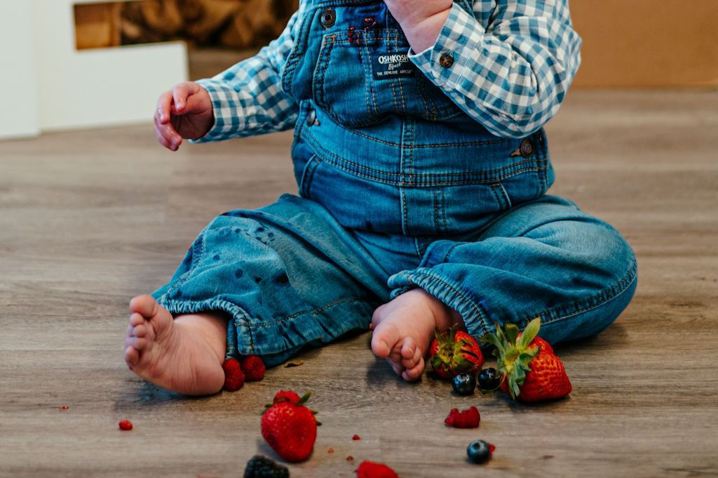 Child in blue denim overalls sitting on the floor with scattered strawberries and berries nearby