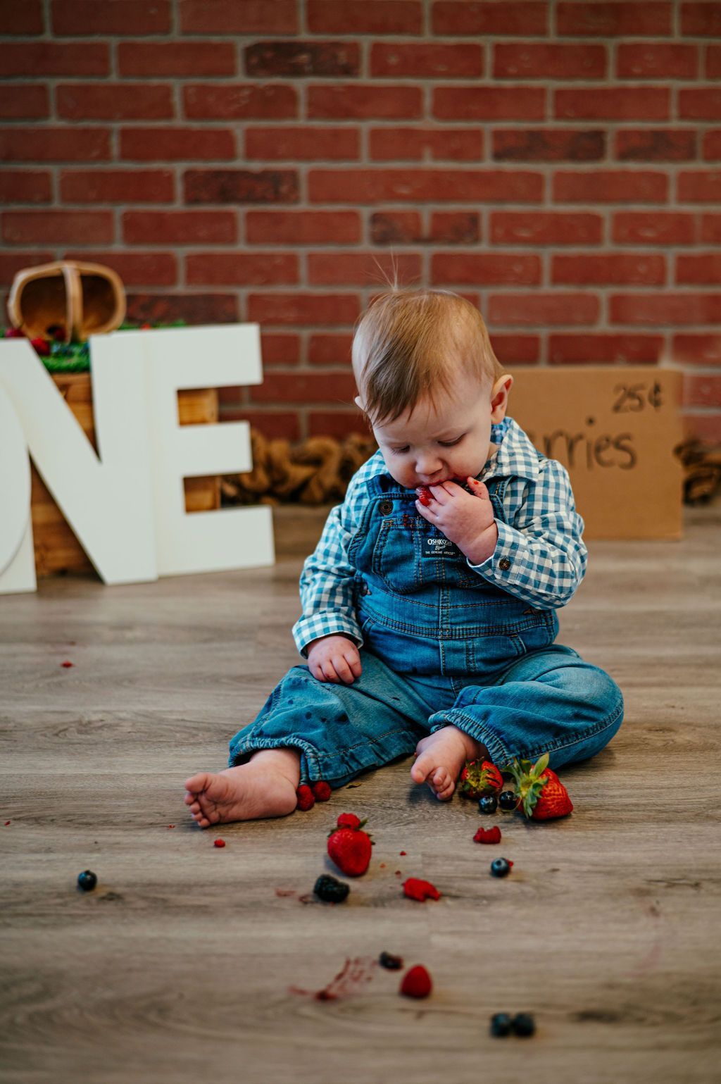 Toddler in blue overalls eating berries on a wooden floor, with a brick wall and large white letters behind.