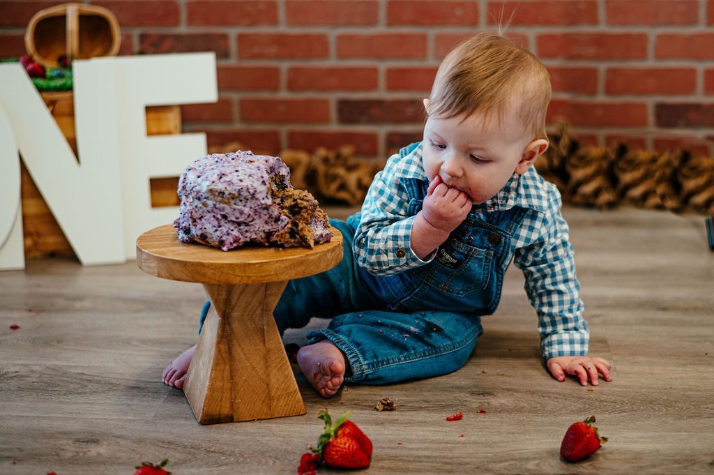 Toddler in blue outfit eating cake on the floor beside a small wooden stand with strawberries and crumbs.