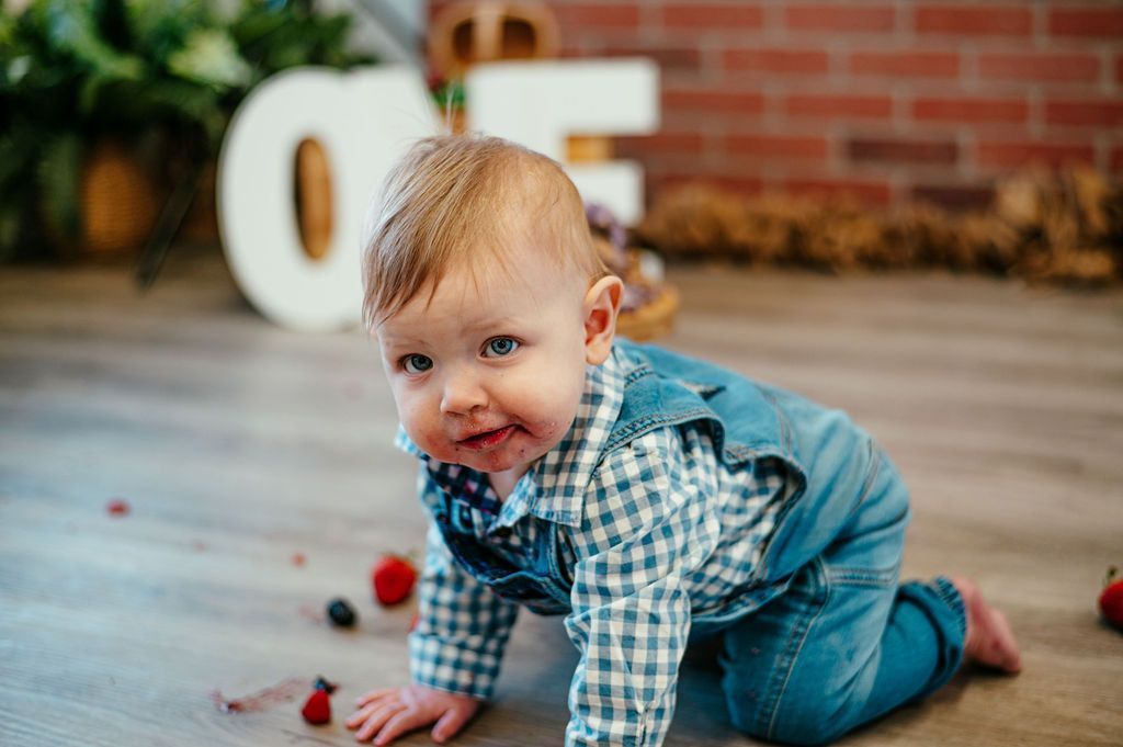 Crawling baby in blue overalls on a wooden floor with scattered berries and a blurred garden backdrop