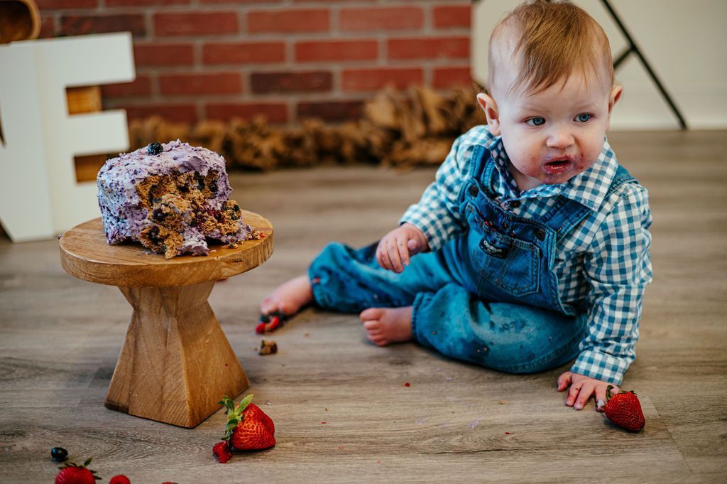 Toddler on the floor reaching toward a messy cake on a small wooden stand, with strawberries nearby.