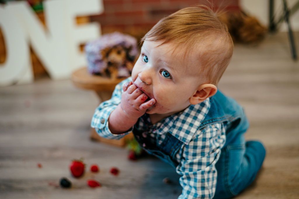 Baby crawling indoors, chewing on fingers beside scattered red berries, wearing a blue checkered shirt.