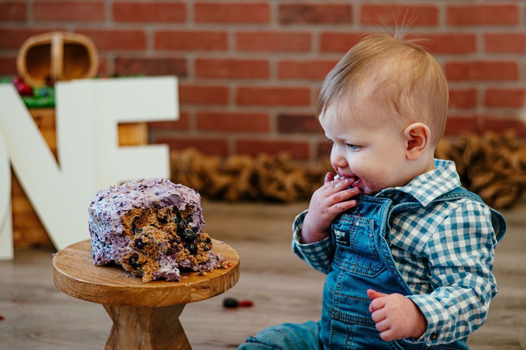 Toddler in blue plaid shirt reaching toward a small frosted cake on a stand, in front of a brick wall and letters.