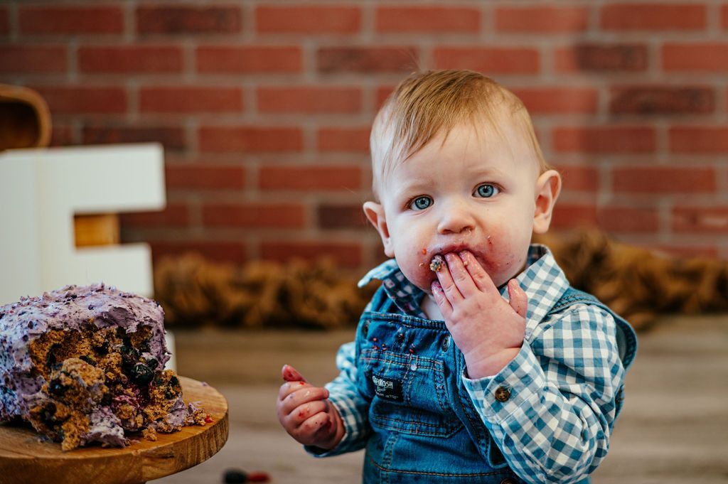 Toddler in blue plaid overalls eating cake at a table, with a brick wall background.