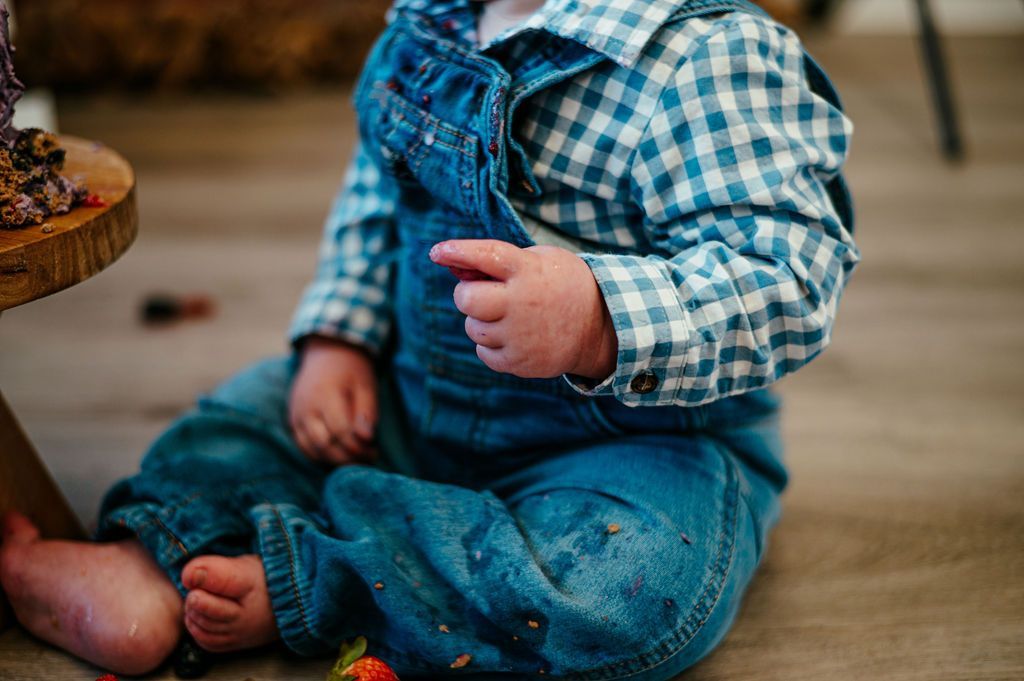 Baby in blue plaid shirt and denim overalls sitting on the floor, holding a small object