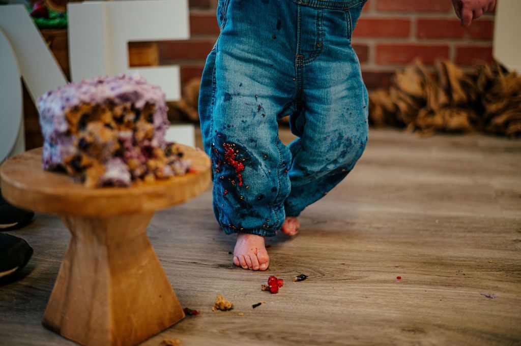 Child’s bare feet covered in cake crumbs beside a small cake on a wooden stand at a party table