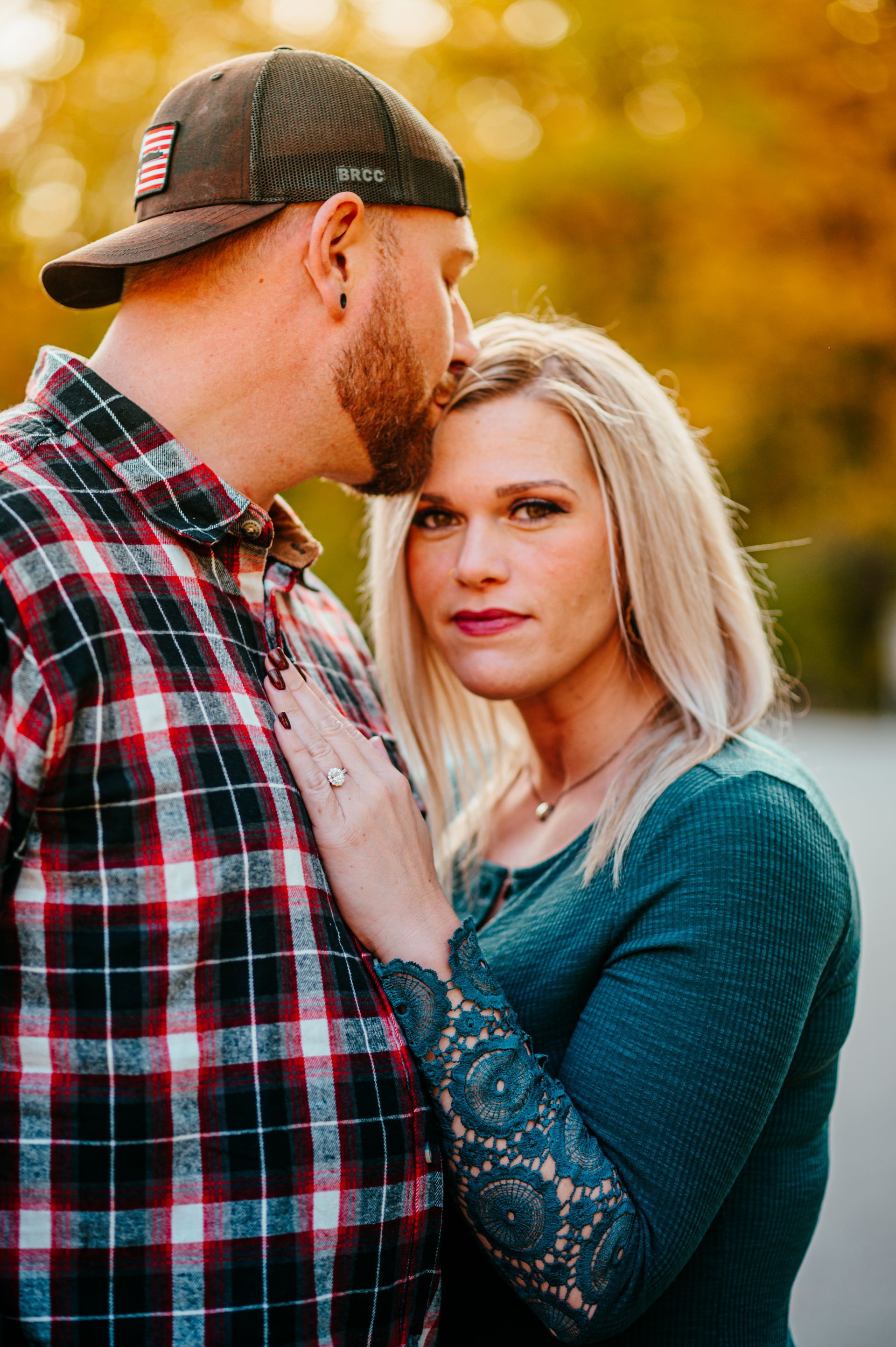 Couple embracing outdoors in warm autumn light, man in plaid shirt and cap, woman in teal sweater.