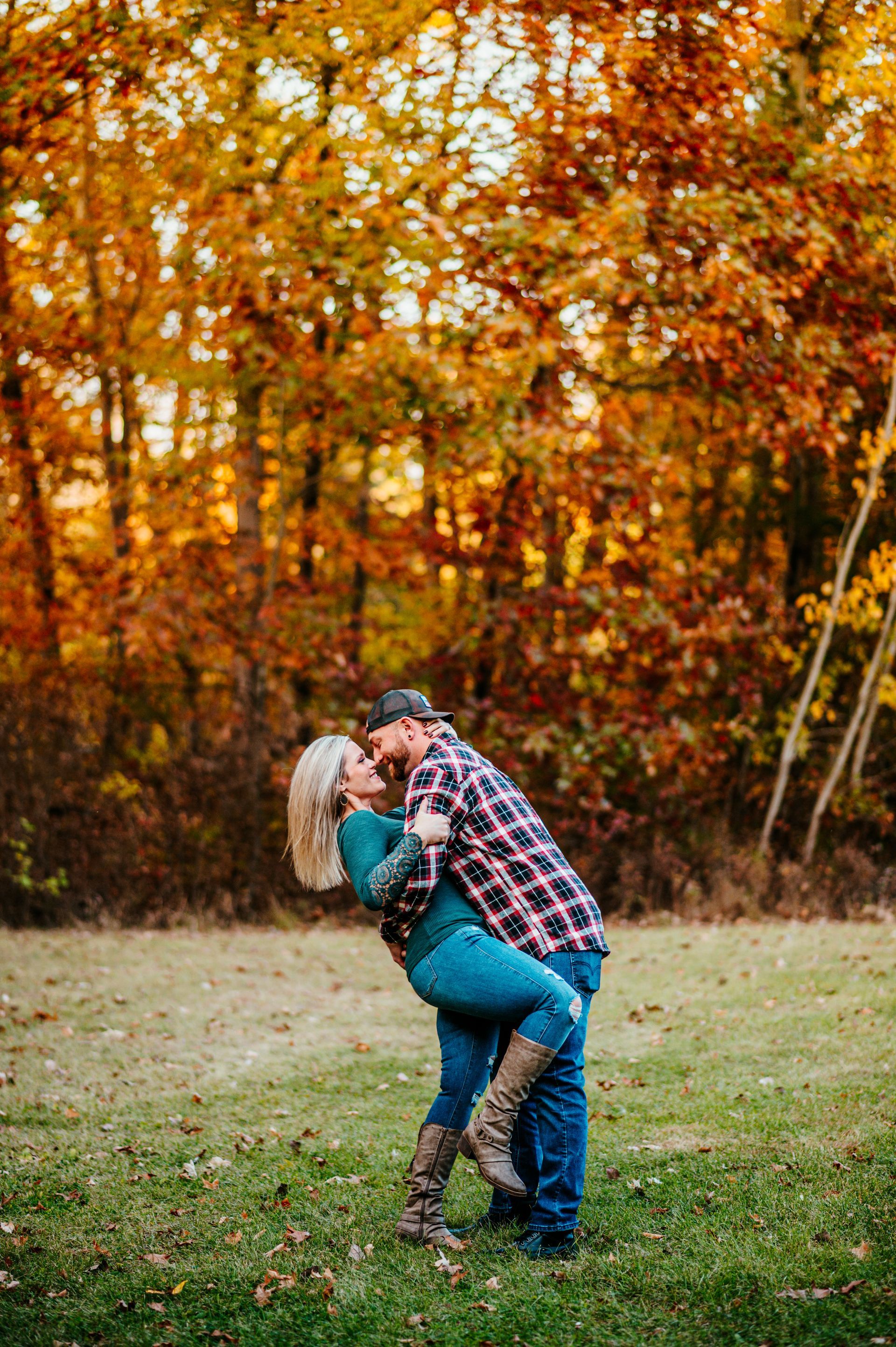 Couple embracing and kissing in a grassy field with autumn trees in the background