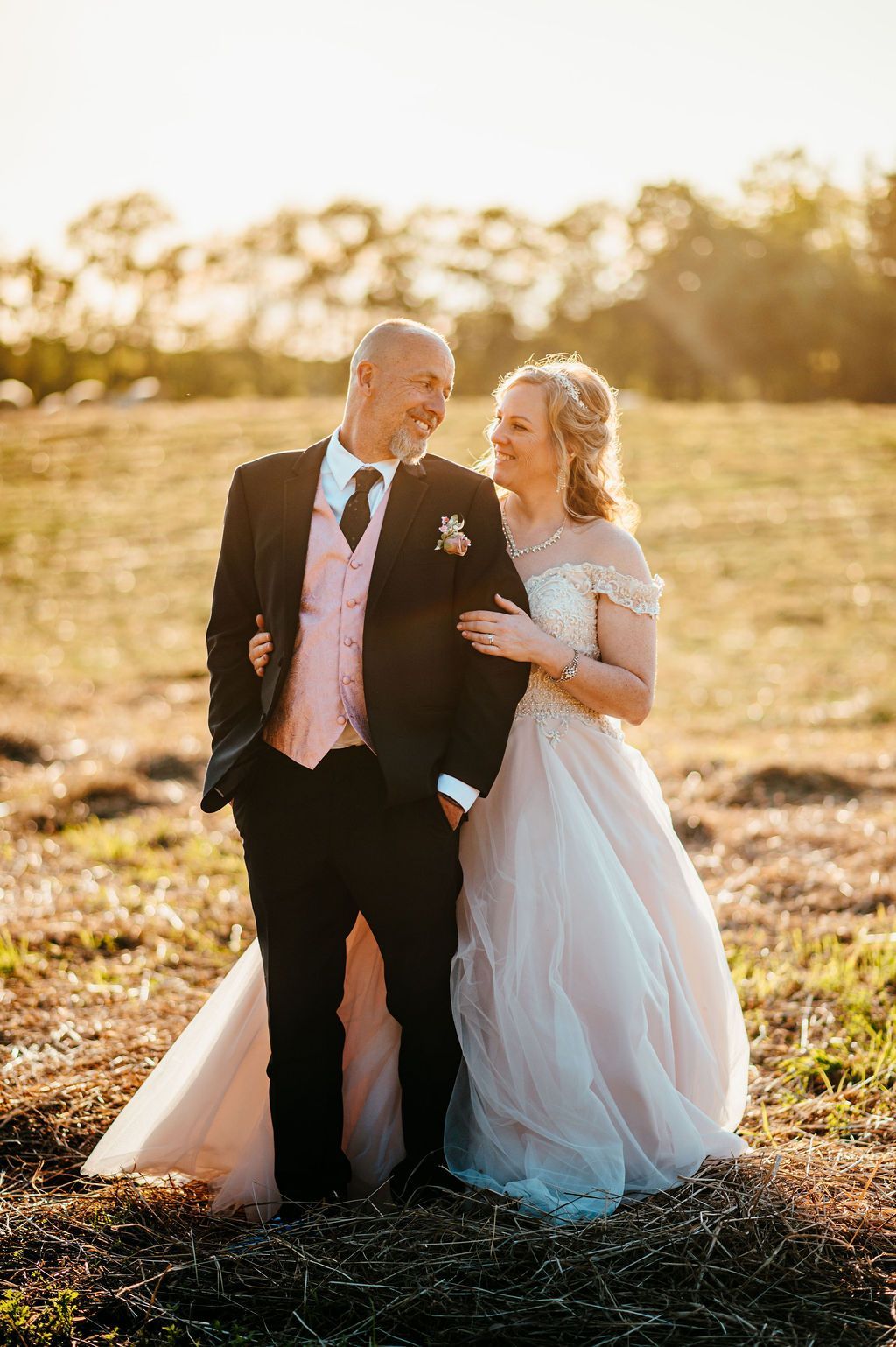 A couple in formal wedding attire walking through a sunlit field at sunset.