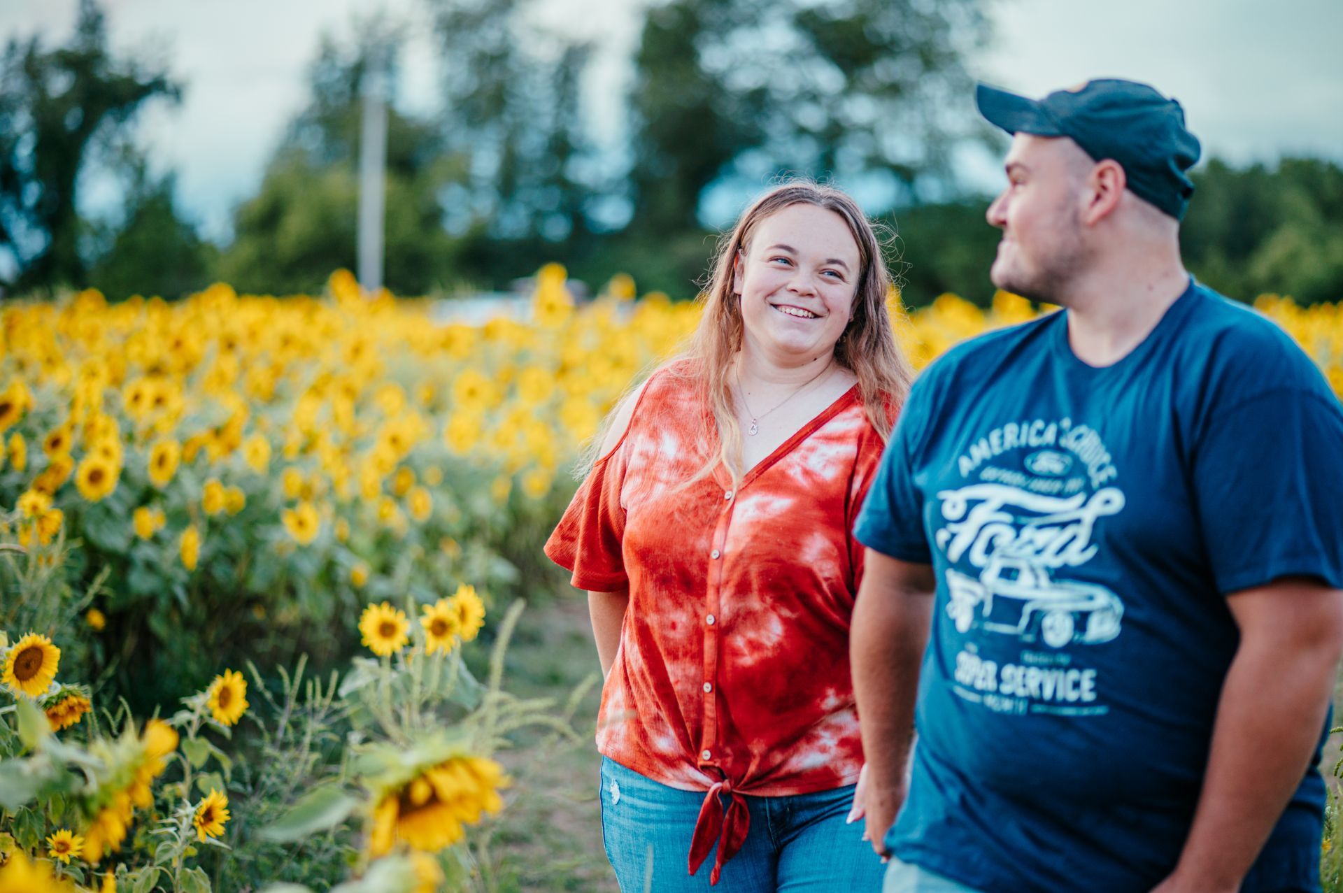Two smiling people walking through a sunflower field at sunset, one in a red tie-dye shirt and one in a blue cap and T-shirt