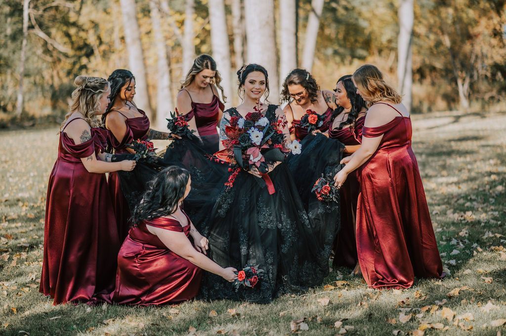 Group in burgundy dresses gathers around a bride in a black gown outdoors, holding red bouquets.
