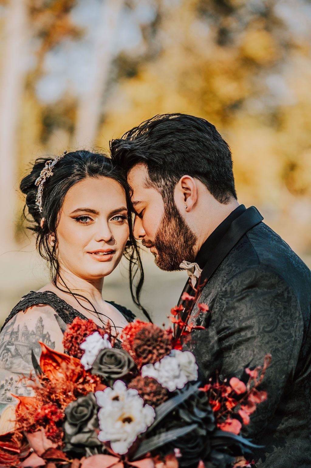 Couple in black outfits holding a red-and-white bouquet outdoors with warm autumn foliage behind them