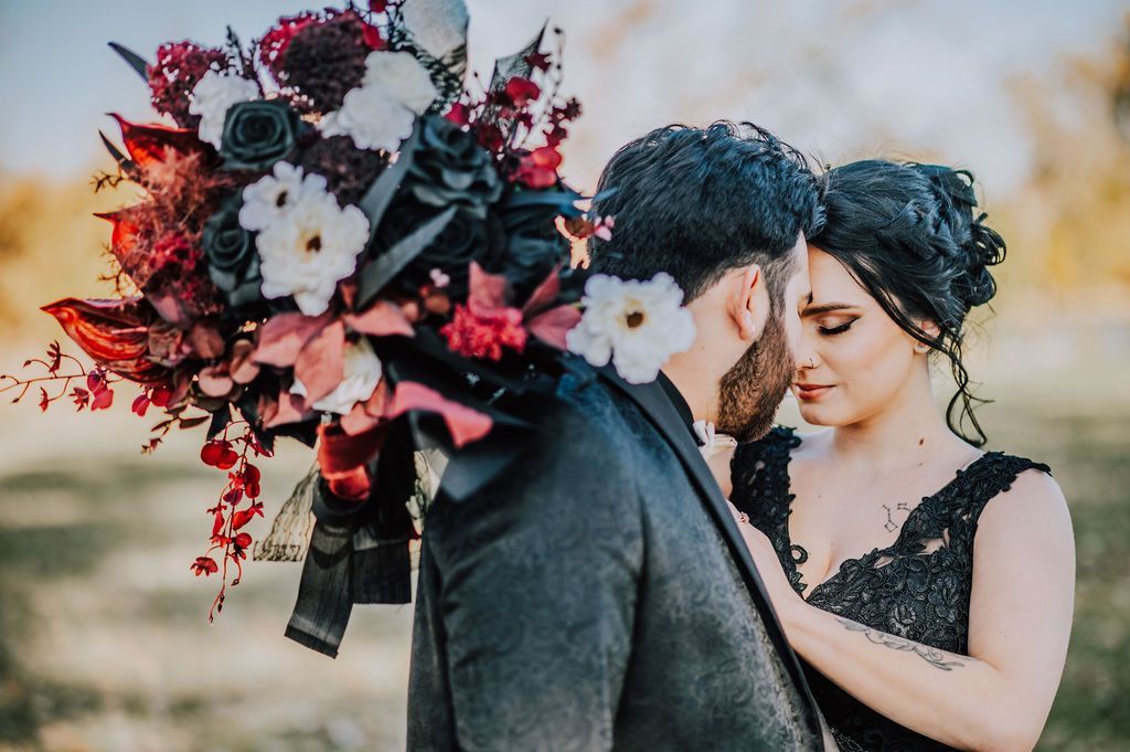 Couple in black embracing outdoors, with a bouquet of red and white flowers behind them.