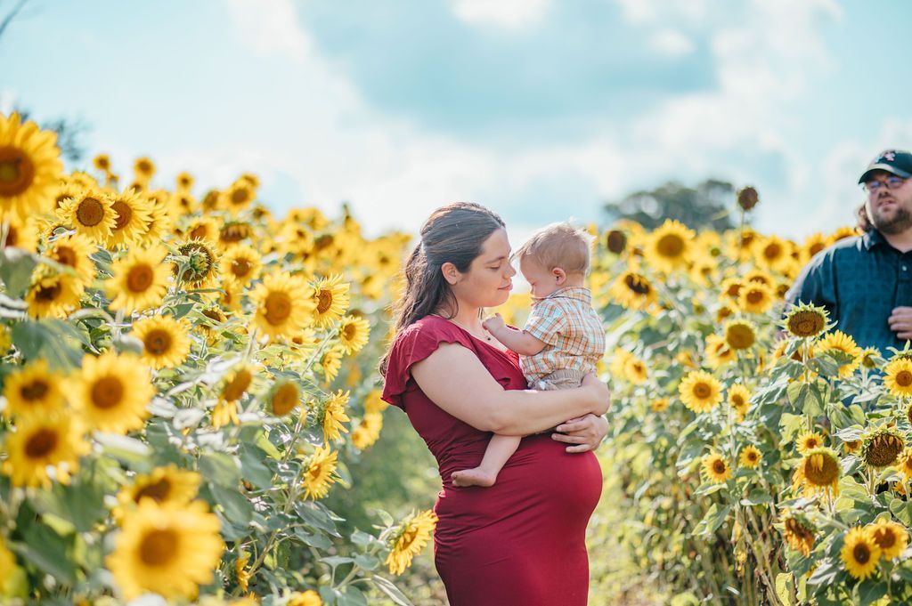 Couple holding a baby in a sunflower field, with a man standing nearby under a bright sky