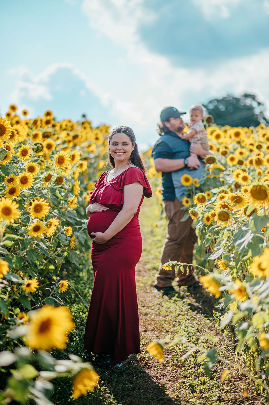 Pregnant woman in a red dress standing in a sunflower field, with a man holding a child behind her