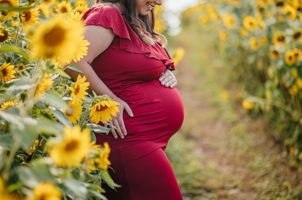 Pregnant person in a red dress standing in a sunflower field with blurred flowers in the foreground