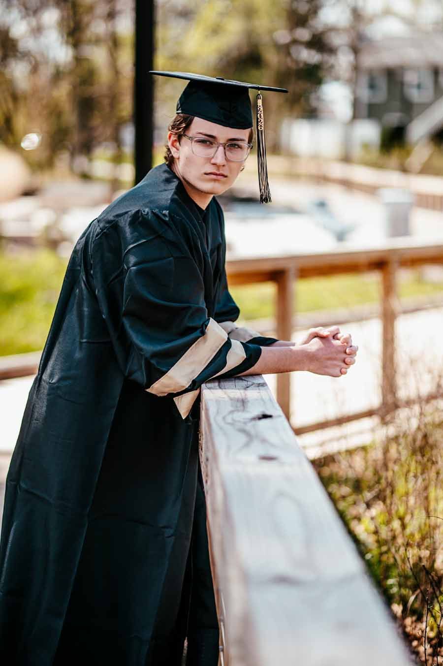 Graduate in black cap and gown leaning on a railing outdoors, looking at the camera.