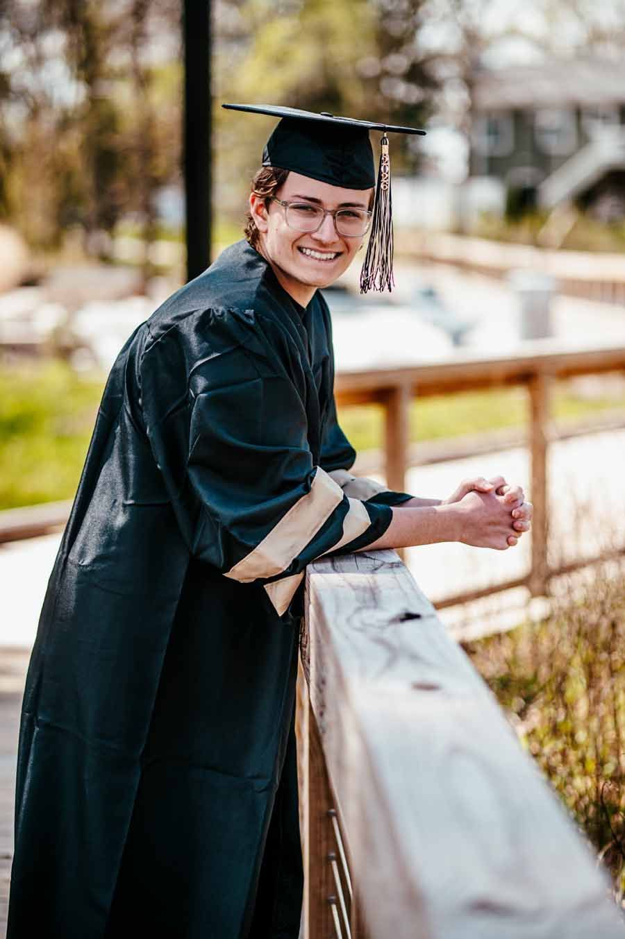 Graduate in black cap and gown smiling while leaning on a wooden railing outdoors