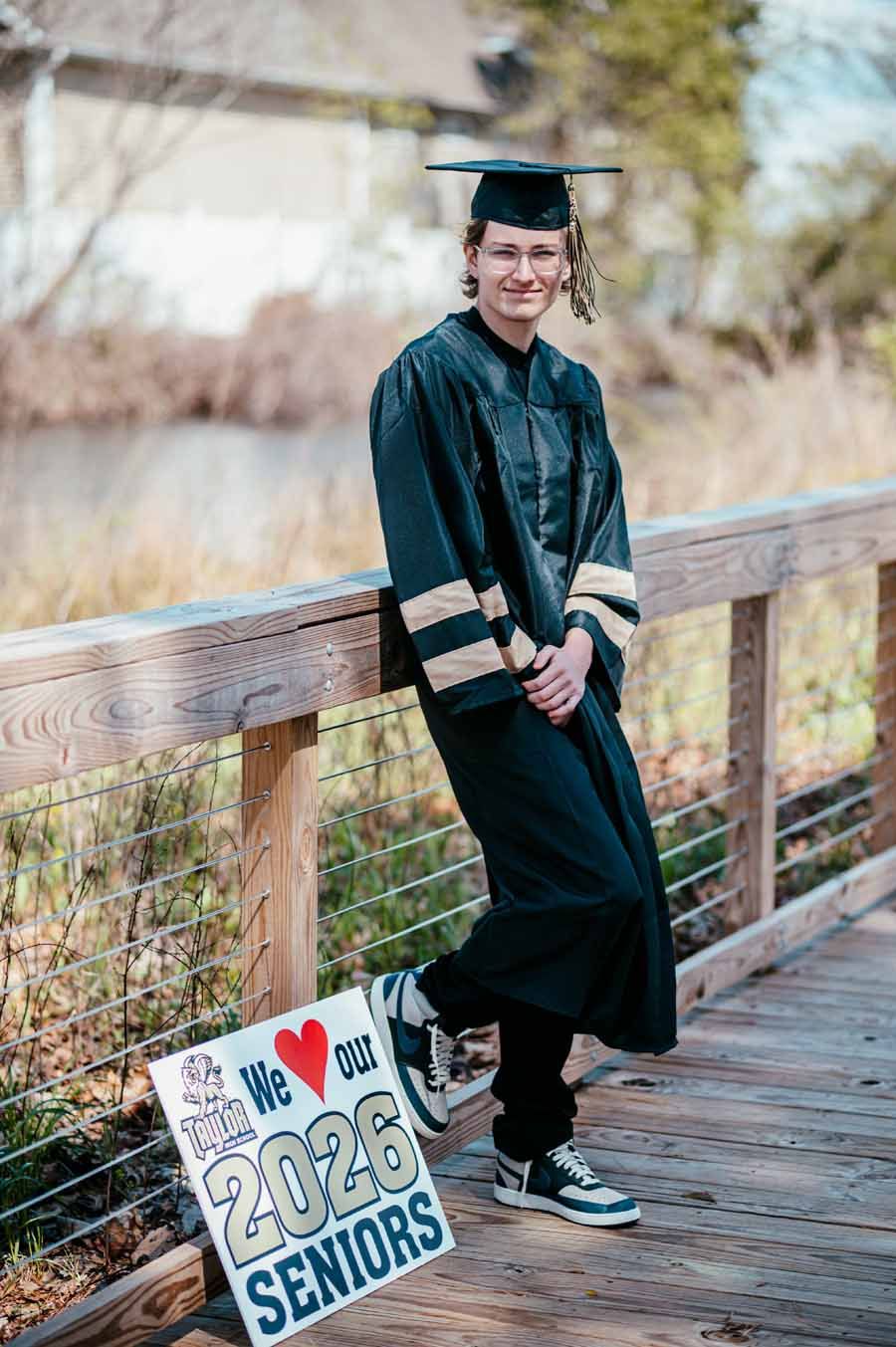 Graduate in cap and gown leaning on a wooden bridge railing beside a “2024 Seniors” sign.