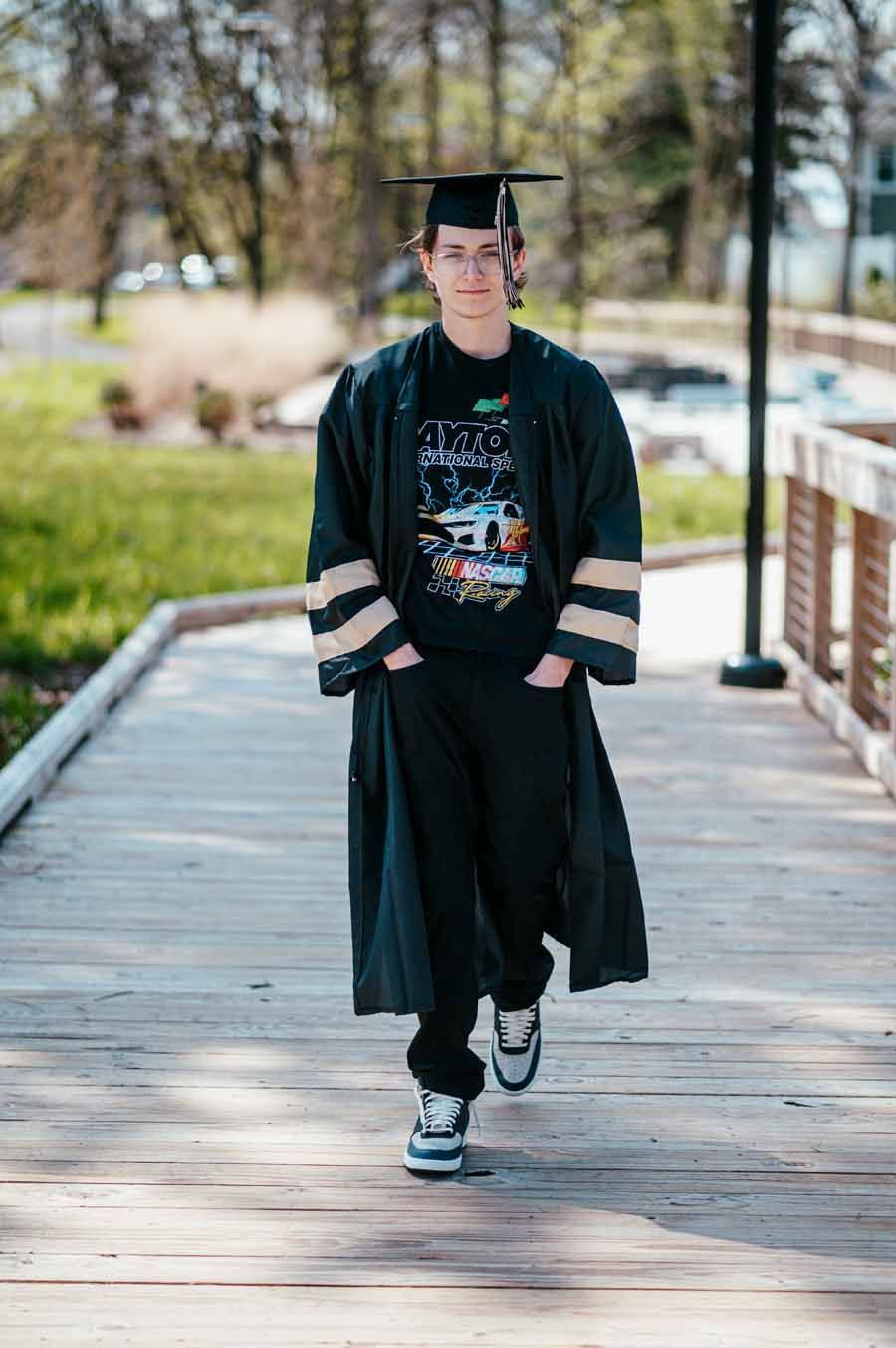 Graduate in cap and gown walking on a wooden bridge outdoors, wearing a black shirt and sneakers.