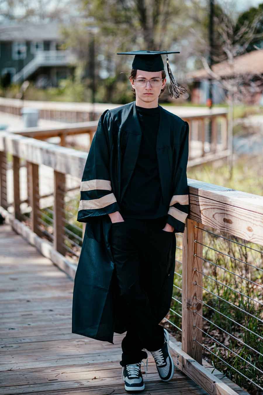 Person in black graduation cap and gown standing on a wooden bridge outdoors.