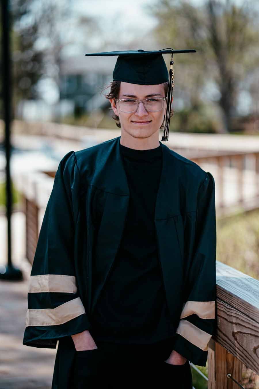 Person in graduation cap and gown standing outdoors on a wooden deck, smiling at the camera.