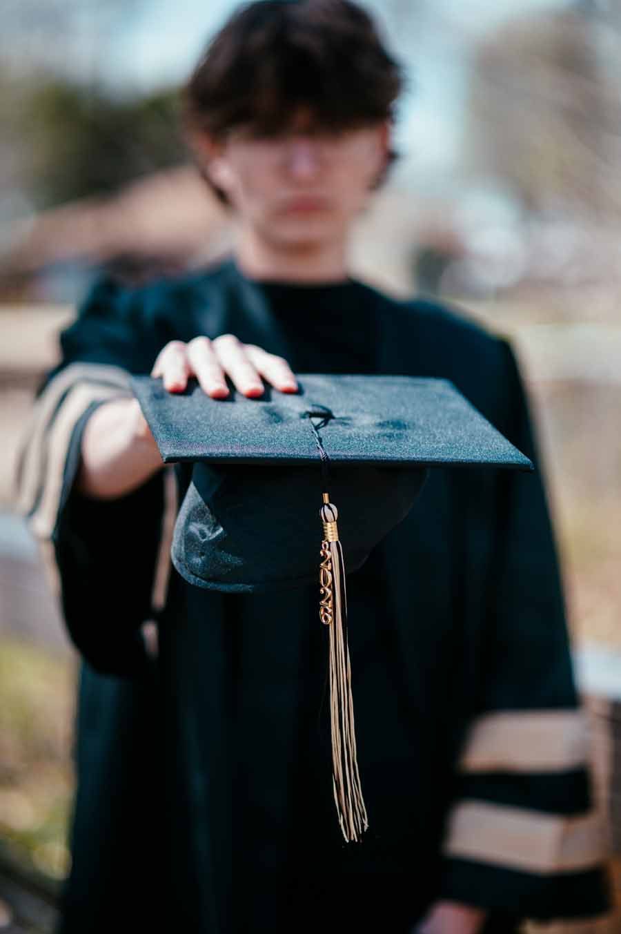 Person in dark academic gown holding out a black graduation cap with tassel.