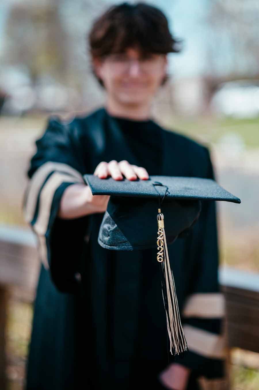 Graduate holding out a black mortarboard with gold tassel outdoors