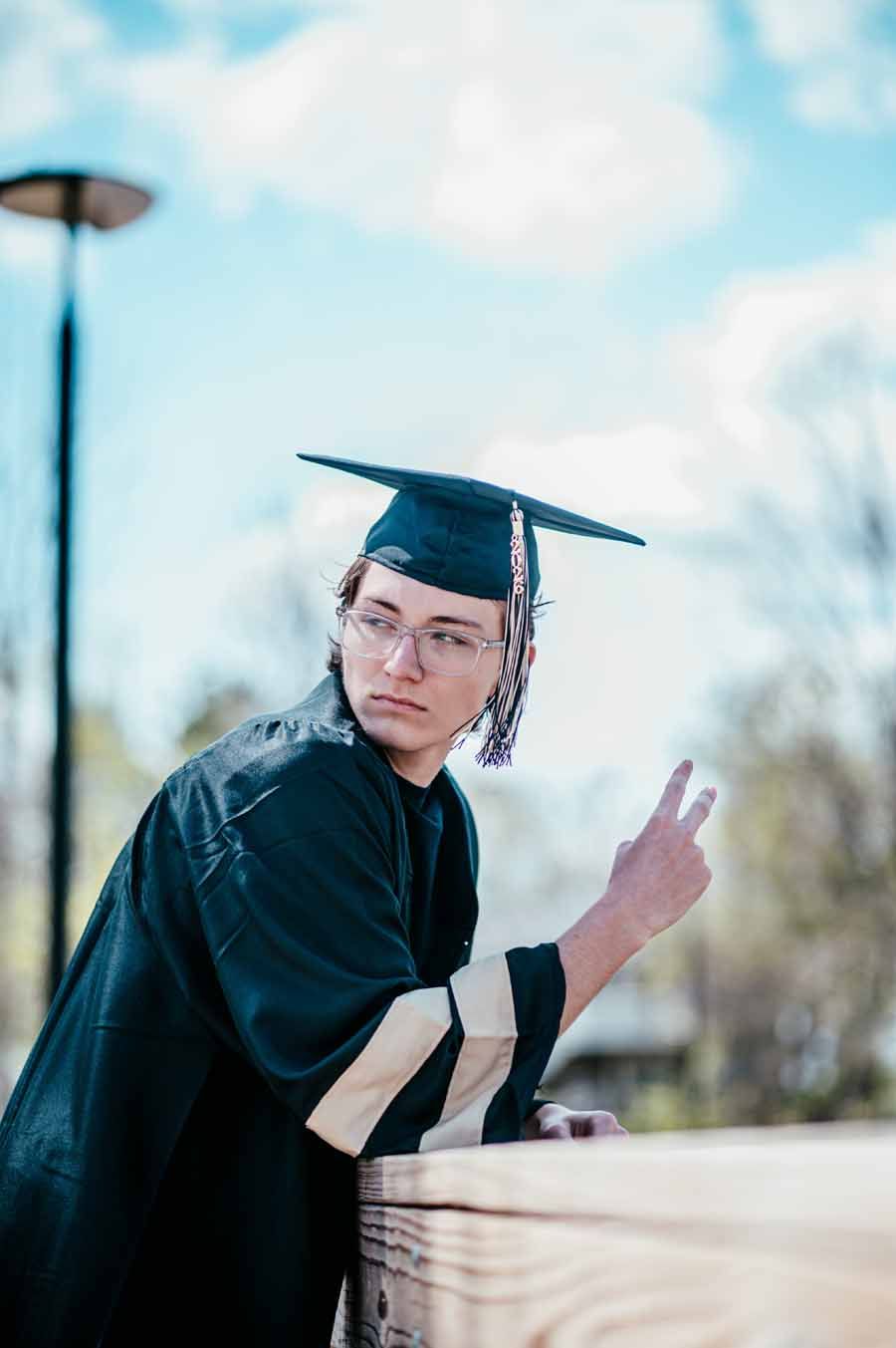 Graduate in cap and gown posing outdoors, making a peace sign beside a blurred tree-lined background