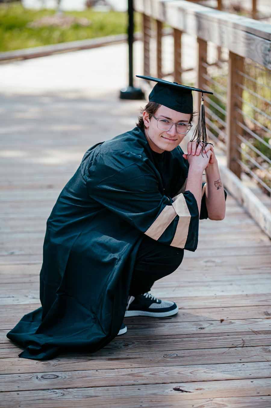 Graduate in black cap and gown crouches on a wooden walkway holding a diploma, smiling at the camera