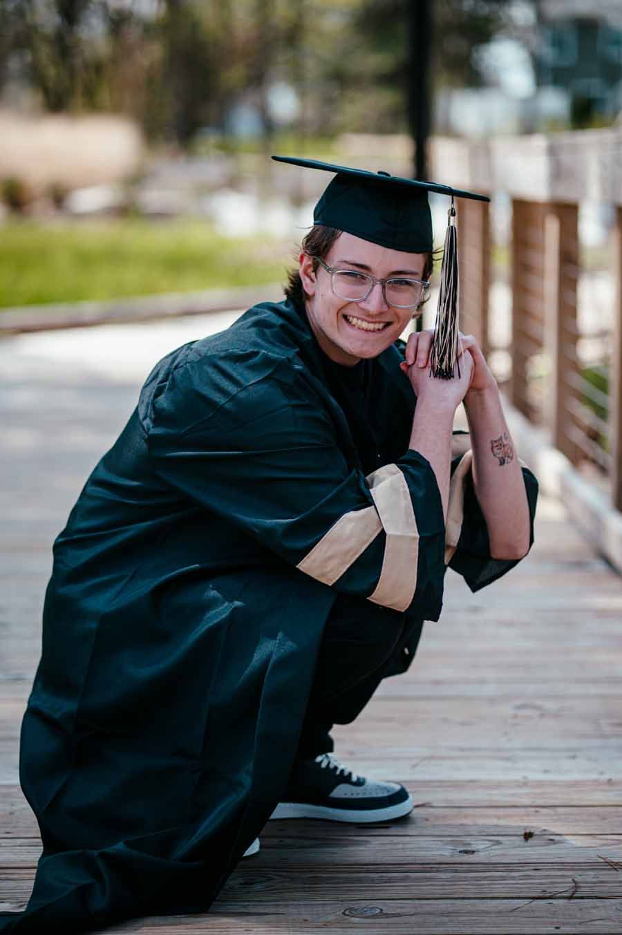 Graduate in black cap and gown crouching on a wooden boardwalk, smiling and holding a diploma.