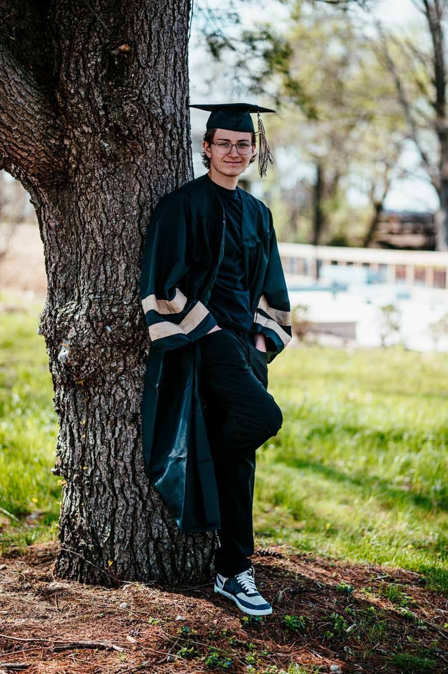 Graduate in cap and gown leaning against a tree outdoors