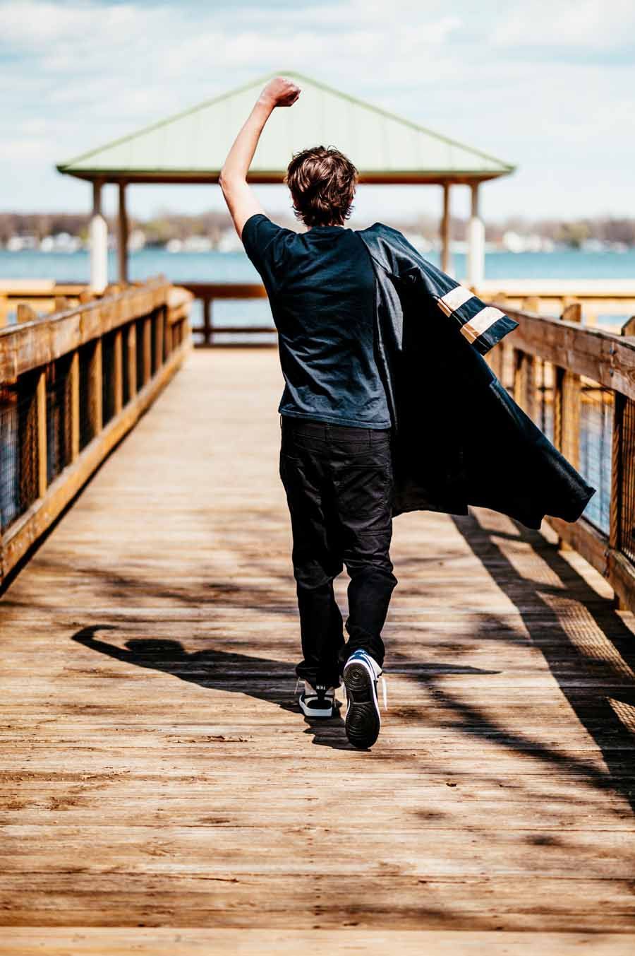 Person walking on a wooden pier, raising one arm, with a gazebo ahead over the water.
