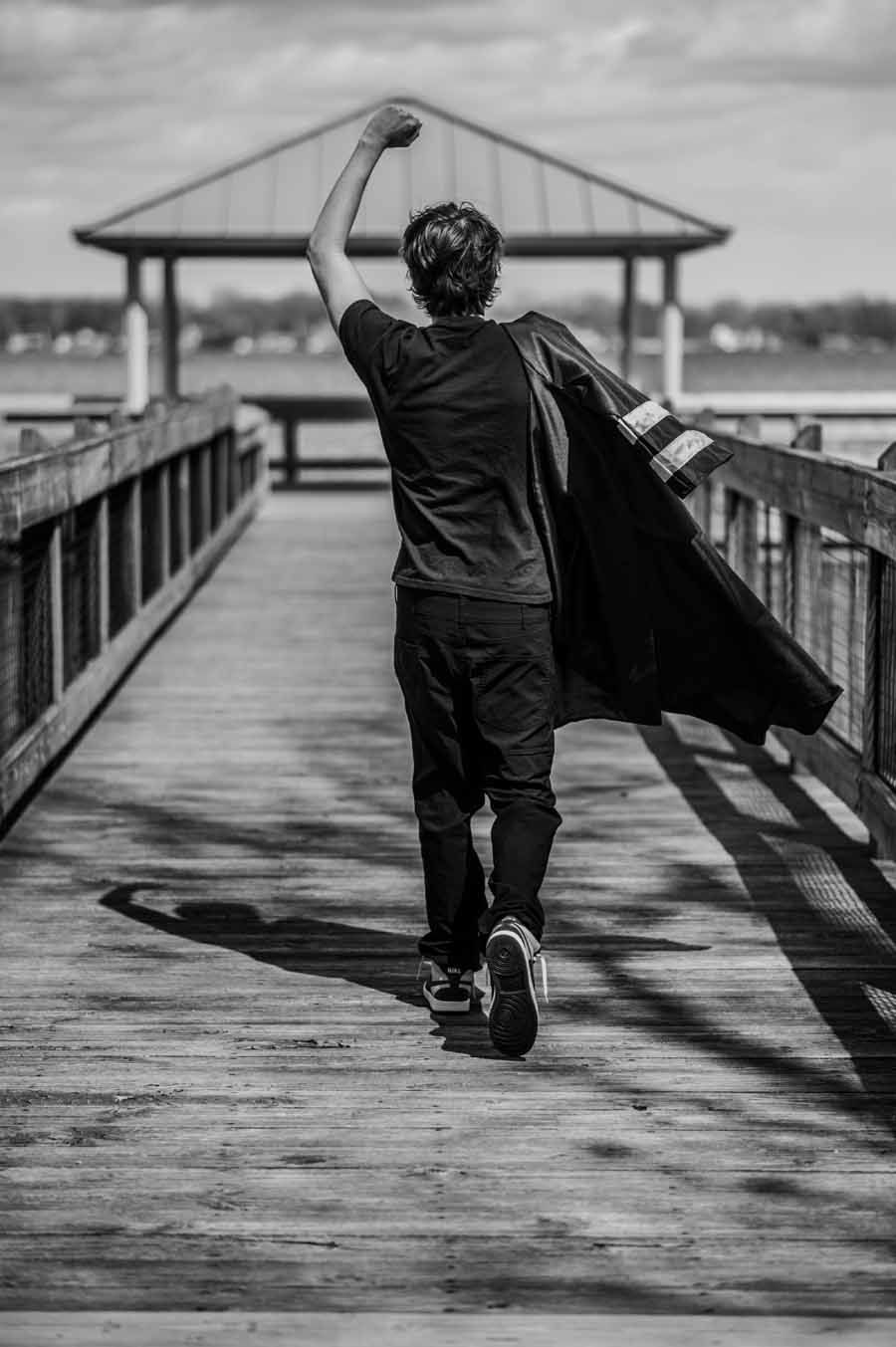 Person walking down a wooden pier with arms raised, wearing dark clothes and carrying a coat or cape.