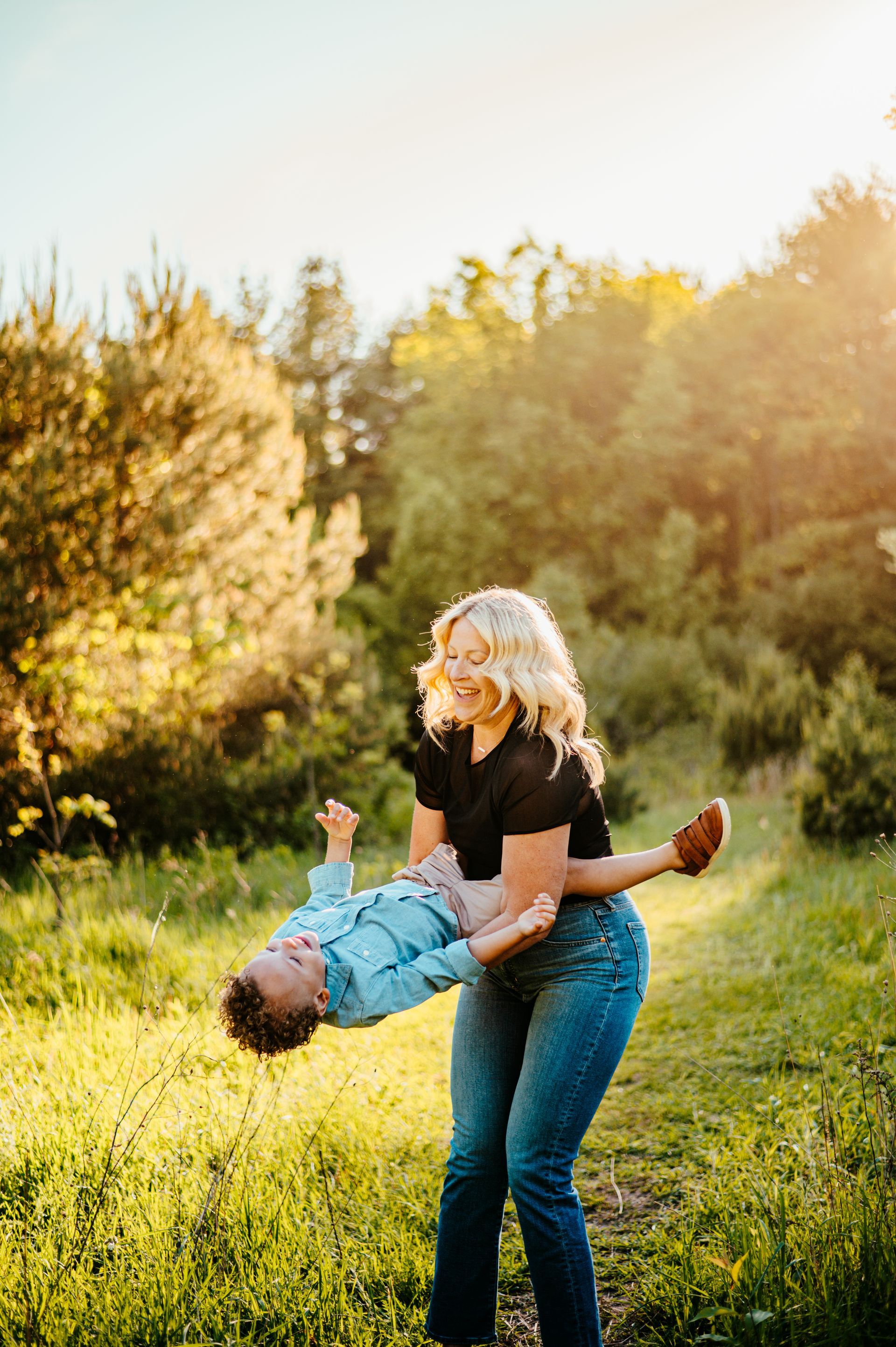 Woman in jeans lifts a smiling child upside down in a sunlit grassy field with trees.