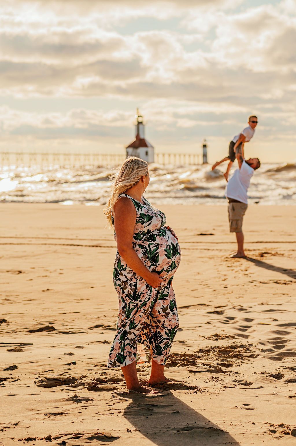 Pregnant person on a beach at sunset, with a man holding a child in the background