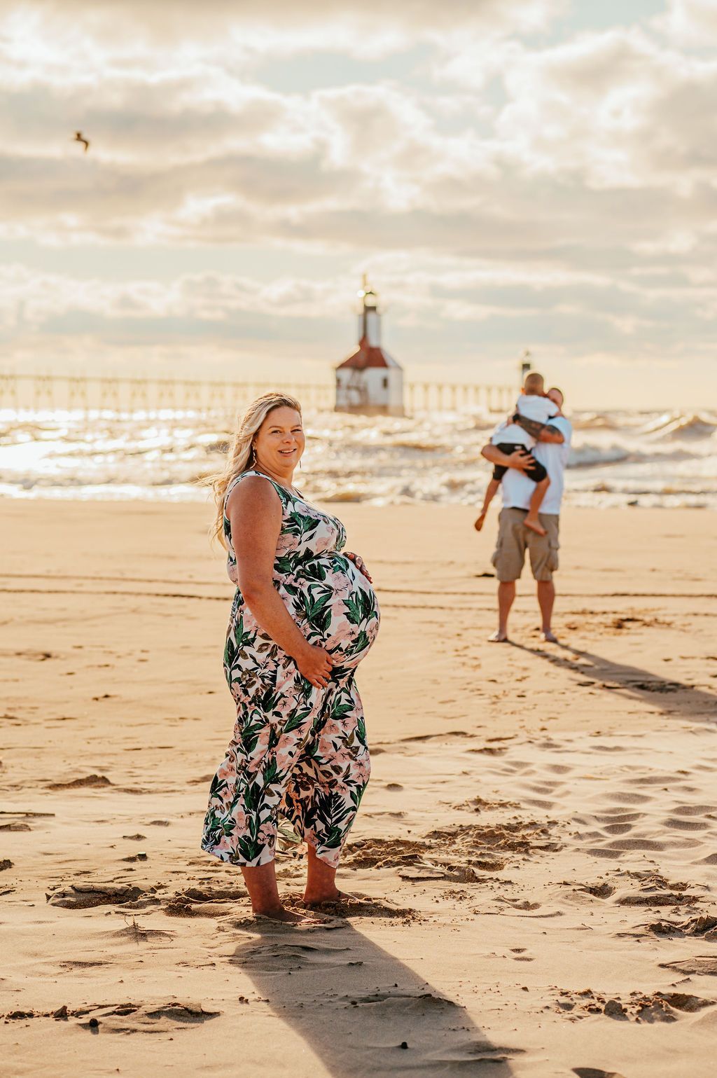 Pregnant person standing on a beach at sunset, with a man carrying a child in the background.