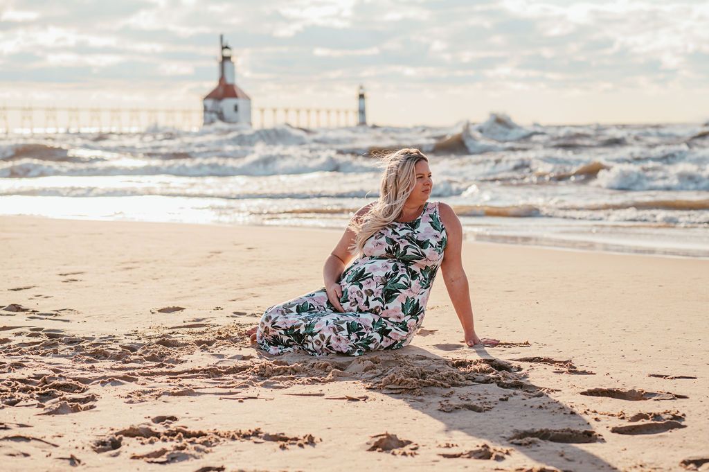 Woman in floral dress sitting on a sandy beach, facing the ocean with a lighthouse in the distance.