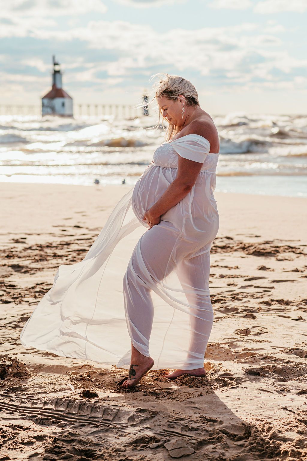 Pregnant person in a sheer white dress walking on a beach at sunset, with a lighthouse in the distance
