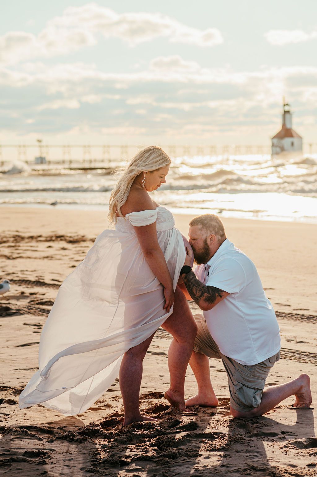 Couple at the beach in soft light, with the man kneeling as he kisses a pregnant belly.